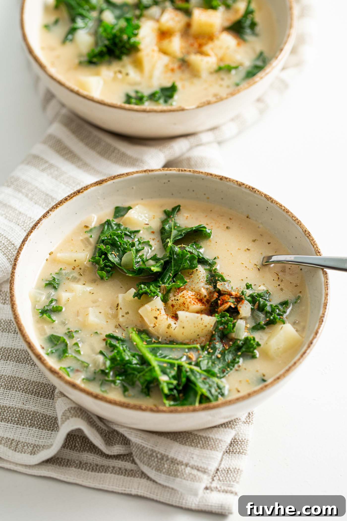 Creamy Plant Based Zuppa Toscana 2 Two steaming bowls of creamy vegan Zuppa Toscana soup, generously filled with tender potato pieces and vibrant green kale, with a spoon gently resting in one bowl, highlighting its inviting texture.