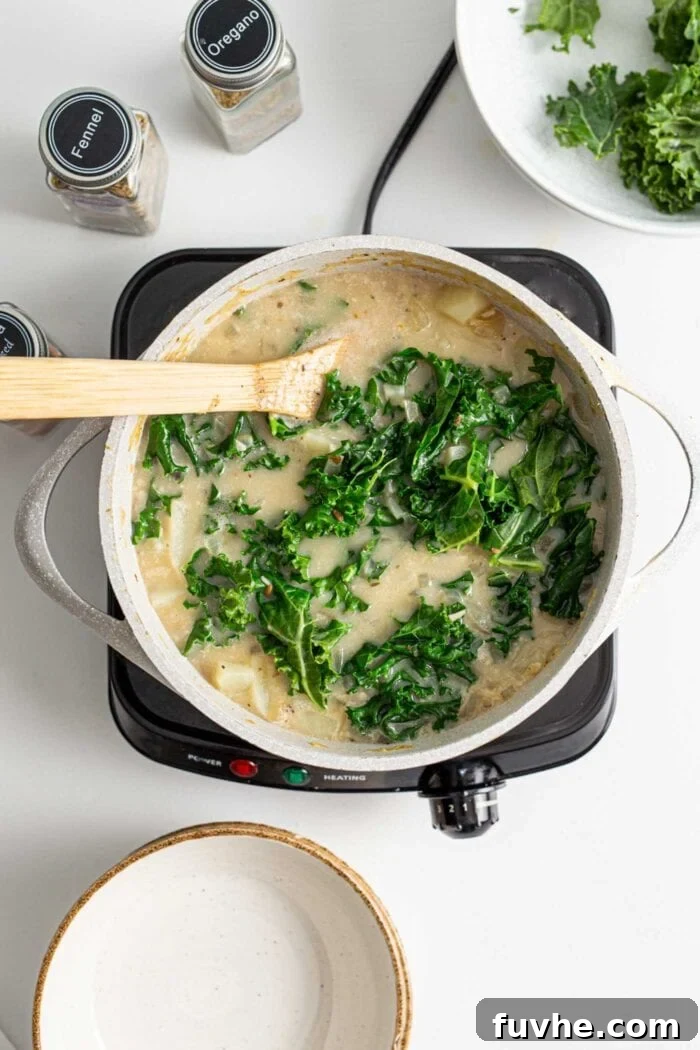 Creamy Plant Based Zuppa Toscana 8 An inviting overhead view of a pot of creamy Zuppa Toscana soup, with vibrant green kale leaves just stirred in, and a wooden spoon resting inside.