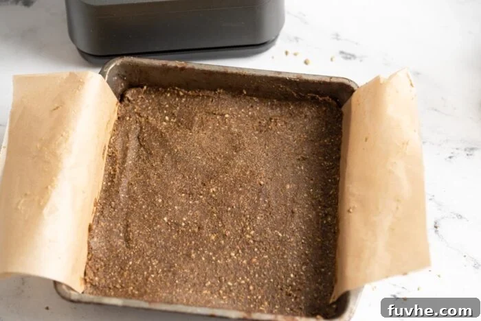 Freshly pressed no-bake energy bar dough evenly spread in a square baking pan lined with parchment paper, ready for chilling and slicing.