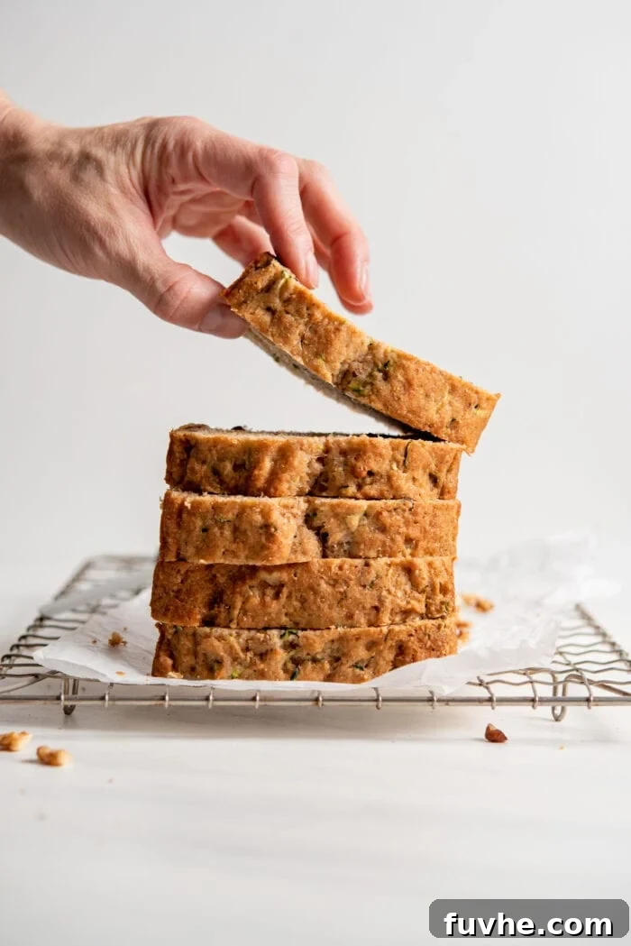 Wholesome Plant-Based Zucchini Bread 14 A hand picking up a slice of zucchini loaf from a stack of slices sitting on a cooling rack.