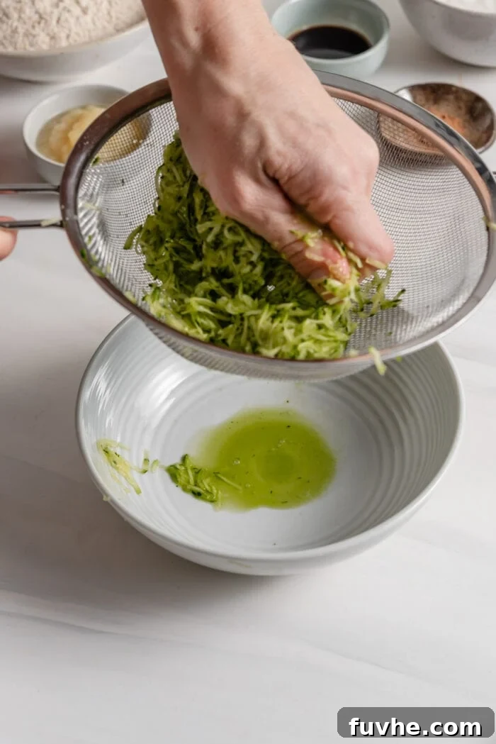 Wholesome Plant-Based Zucchini Bread 5 A hand squeezing grated zucchini over a strainer to demonstrate removing excess water.