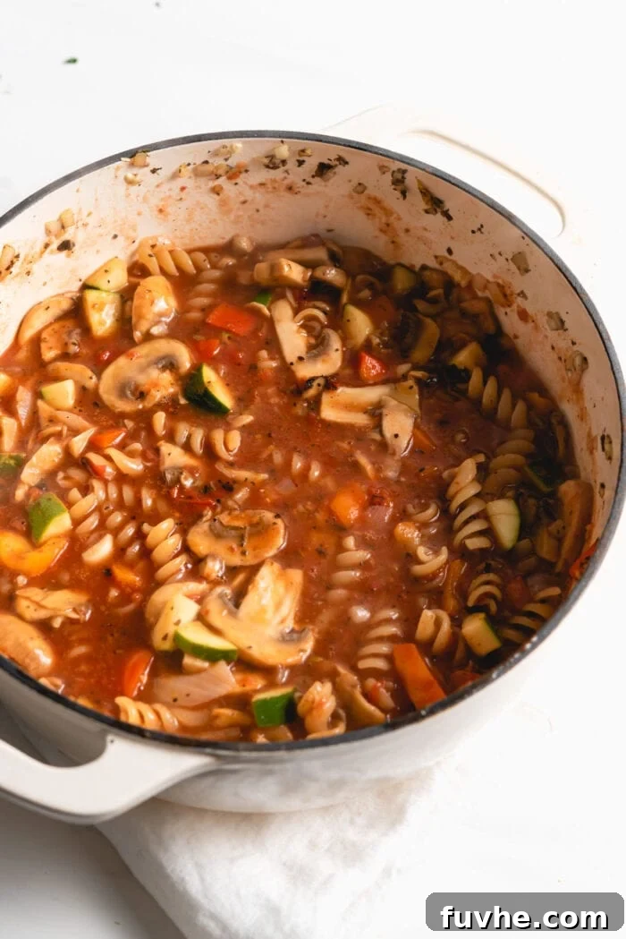 Rotini pasta, mixed with vegetables, submerged in a rich, simmering tomato sauce within the one-pot, beginning to cook.