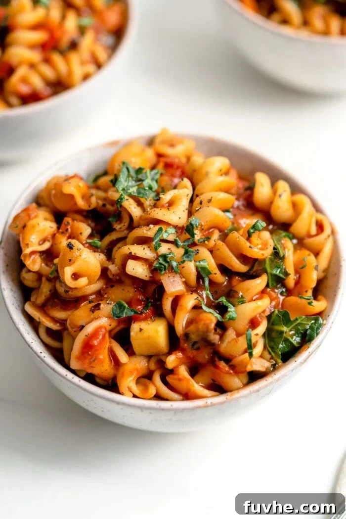 A close-up of a rustic bowl of vegan one-pot pasta, showcasing the perfectly cooked noodles coated in a rich tomato and vegetable sauce, garnished with fresh chopped herbs.