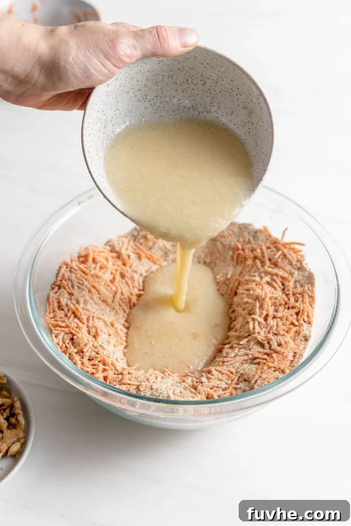 Pouring the combined wet ingredients (applesauce, coconut oil, milk) into a created 'well' in the center of the dry ingredients and grated carrot mixture in a large bowl.