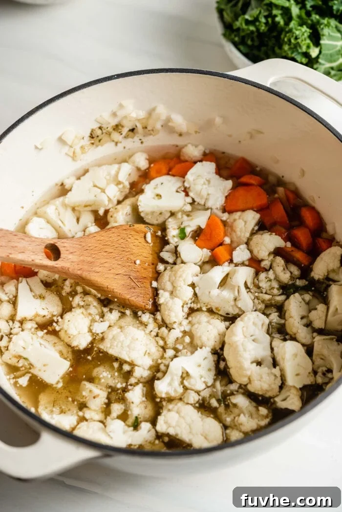 Chopped cauliflower and carrot cooking in a large soup pot with a wooden spoon, simmering in broth.