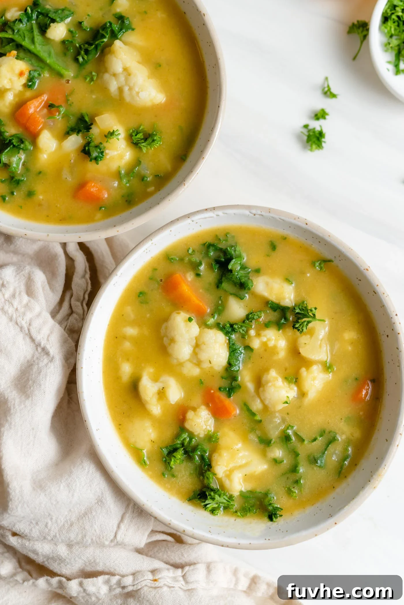 Overhead view of two bowls of vibrant kale, cauliflower, and carrot soup, garnished for serving.