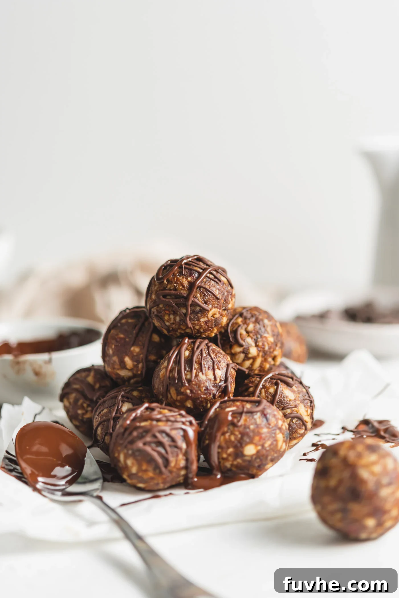 Stack of no-bake energy balls drizzled in chocolate sitting on a folded piece of parchment paper beside a spoon with melted chocolate on it. These vibrant, healthy snacks are perfect for a quick energy boost.