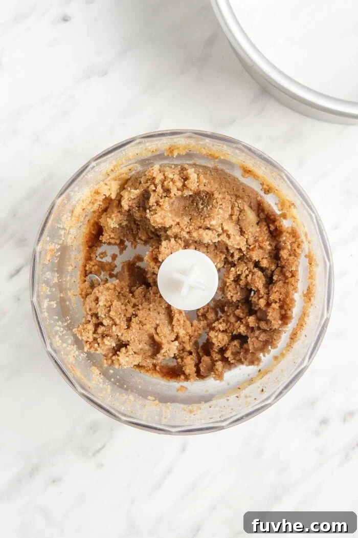 The blended raw pie crust mixture, consisting of almond flour, pecans, maple syrup, and cinnamon, visible within a food processor bowl, ready for pressing.