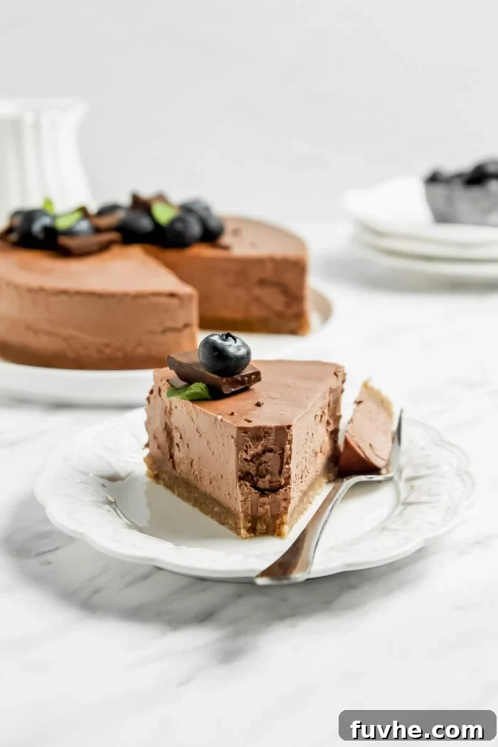 A beautifully presented slice of chocolate cheesecake, adorned with a few blueberries, resting on a small decorative plate with a fork, while a whole cheesecake sits blurred in the background on a cake stand.