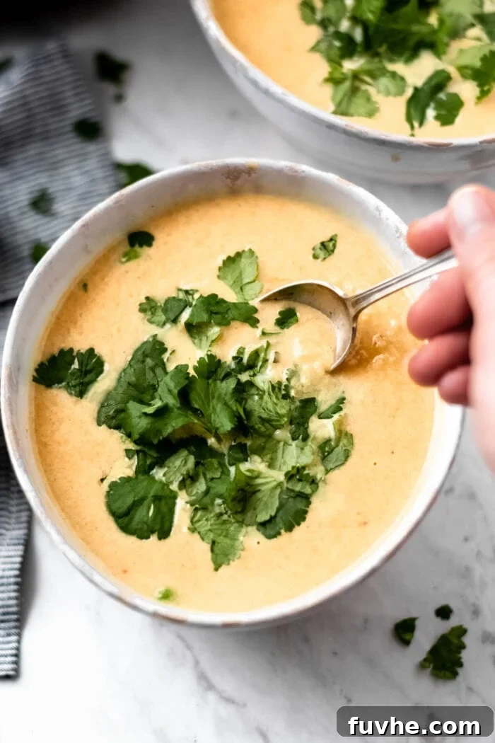 A hand stirring a bowl of creamy cauliflower sweet potato soup topped with fresh cilantro with a spoon.