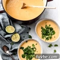 Two bowls and a large stock pot of cauliflower sweet potato soup with green peas. The bowls of soup are topped with fresh cilantro and there are some wedges of lime and two spoons beside them.