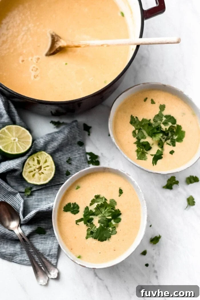 Two bowls and a large stock pot of cauliflower sweet potato soup with green peas. The bowls of soup are topped with fresh cilantro and there are some wedges of lime and two spoons beside them.