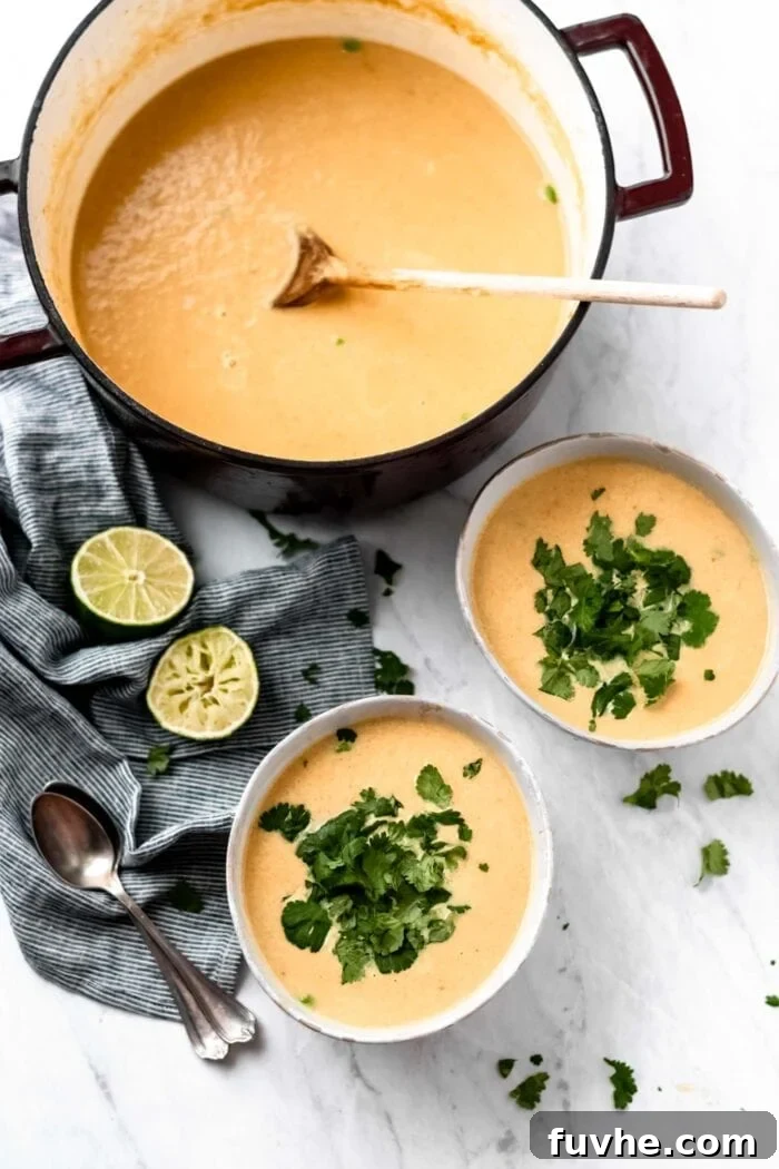Two bowls and a large stock pot of cauliflower sweet potato soup with green peas. The bowls of soup are topped with fresh cilantro and there are some wedges of lime and two spoons beside them.