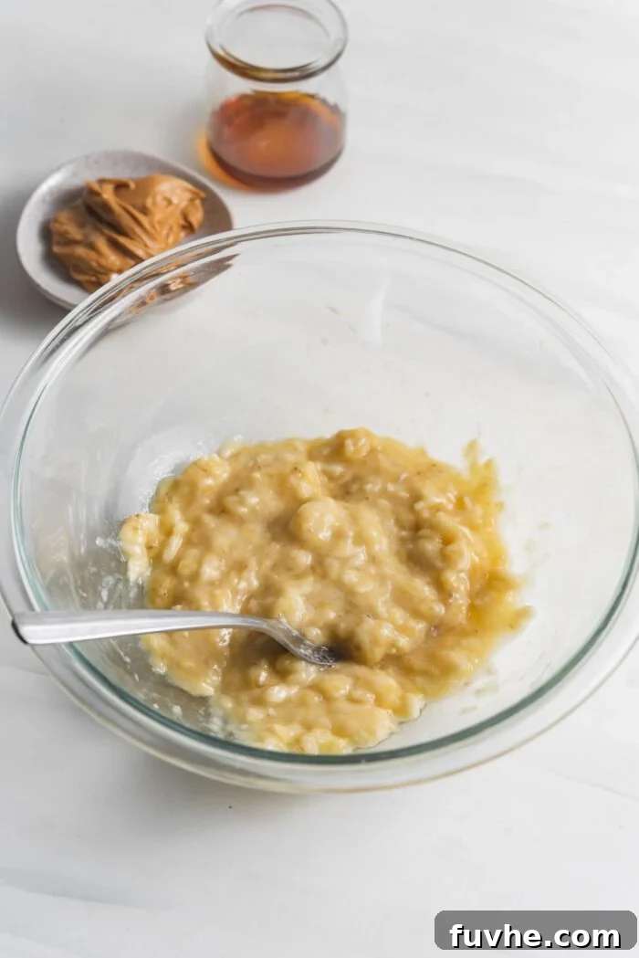 Ripe bananas being mashed into a smooth paste in a clear glass mixing bowl using a fork.