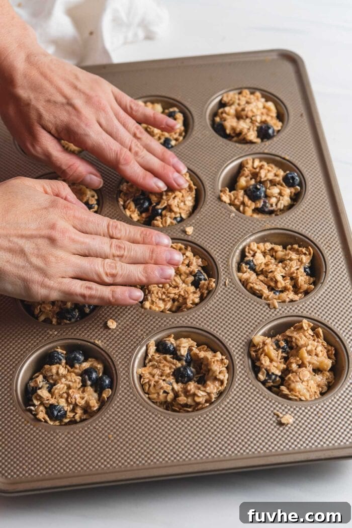 Two hands pressing the oatmeal cup batter firmly into a 12-cup muffin tin, ensuring an even layer.