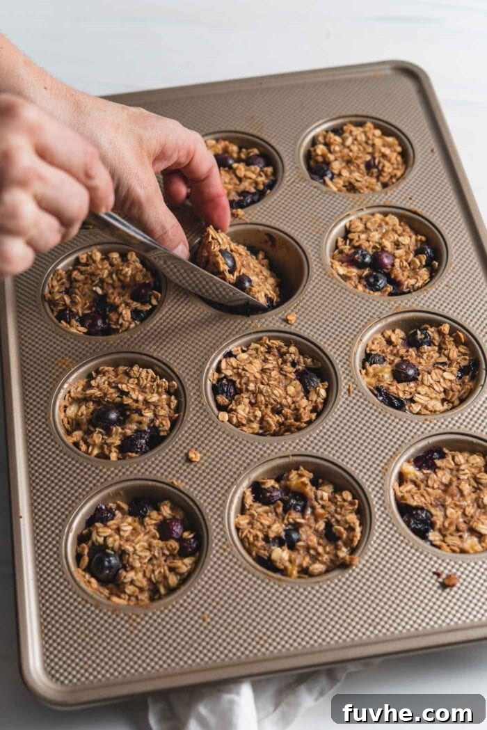 A hand using a knife to carefully loosen and remove a baked oatmeal muffin from a 12-cup muffin pan, showing the golden-brown edges.