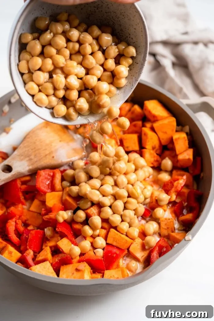 Hearty Sweet Potato Chickpea Coconut Curry 8 A bowl of chickpeas, including their liquid, being poured into a skillet filled with sweet potato and bell pepper, ready to simmer.