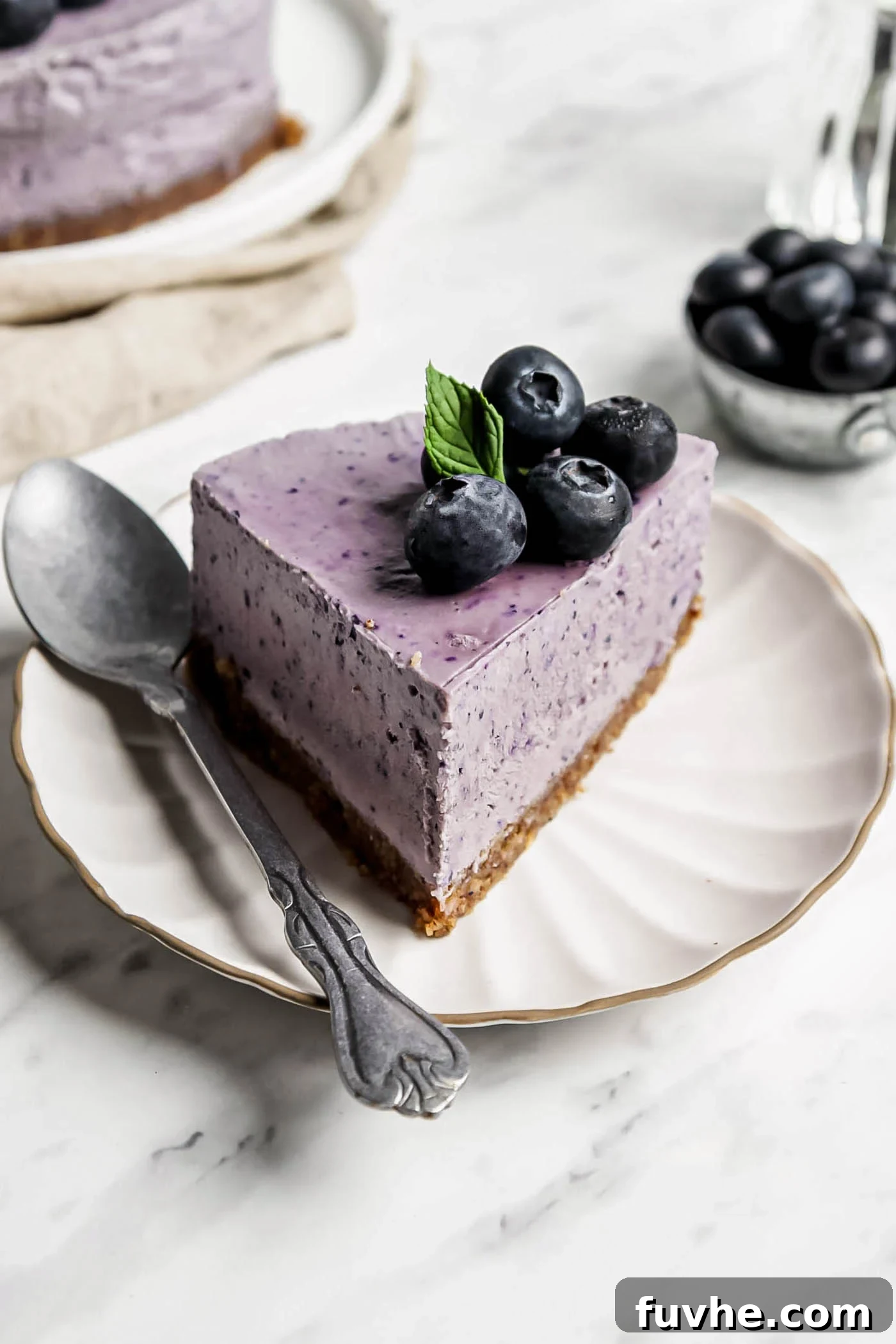 A perfectly sliced piece of vegan blueberry cheesecake, showcasing its vibrant purple filling and golden crust, served on a small plate with a spoon, ready to be enjoyed.