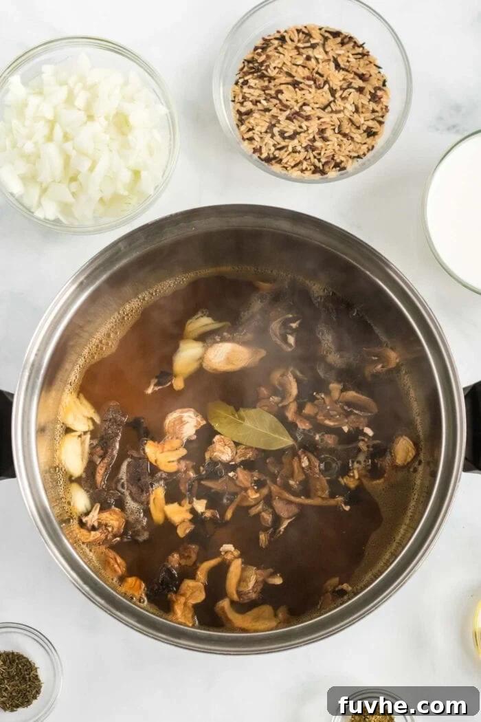 A pot of simmering vegetable broth with mixed dried mushrooms, crushed garlic, and a bay leaf, beginning their rehydration process.