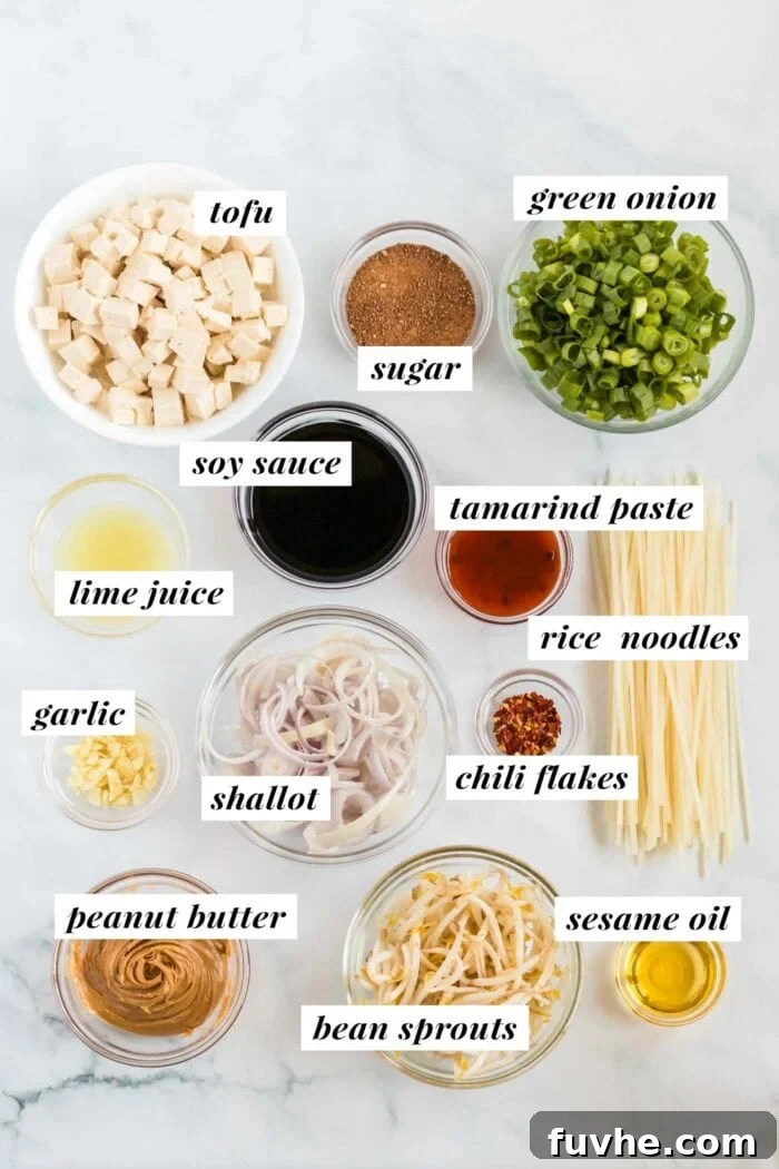 A visual display of all the fresh ingredients required for preparing a vegan tofu Pad Thai, including wide rice noodles, pressed tofu, shallots, green onions, garlic, lime, cilantro, and containers for tamarind paste, soy sauce, and peanut butter.