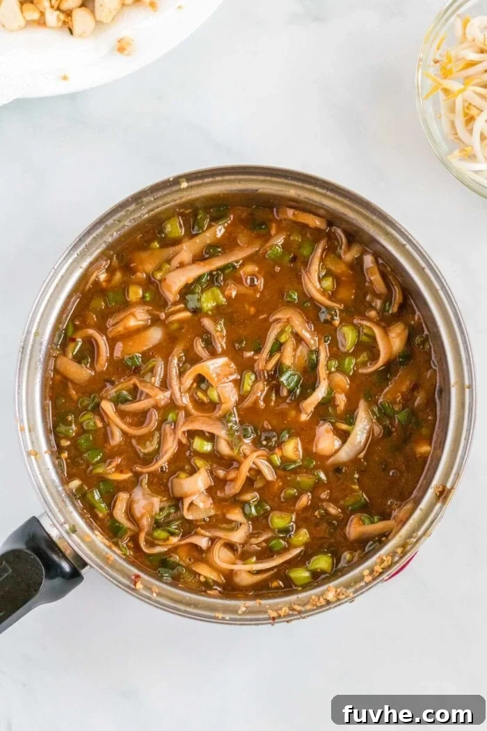 The homemade Pad Thai sauce, now thickened and gently simmering in a pan, infused with the sautéed aromatics like scallions and shallots, creating a rich base for the noodles.