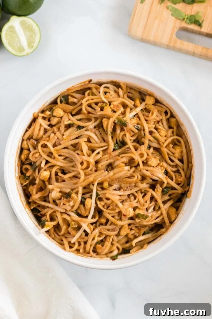 Cooked wide rice noodles being vigorously tossed with the rich, simmering Pad Thai sauce in a large pan using tongs, ensuring every strand is thoroughly coated in the flavorful sauce.