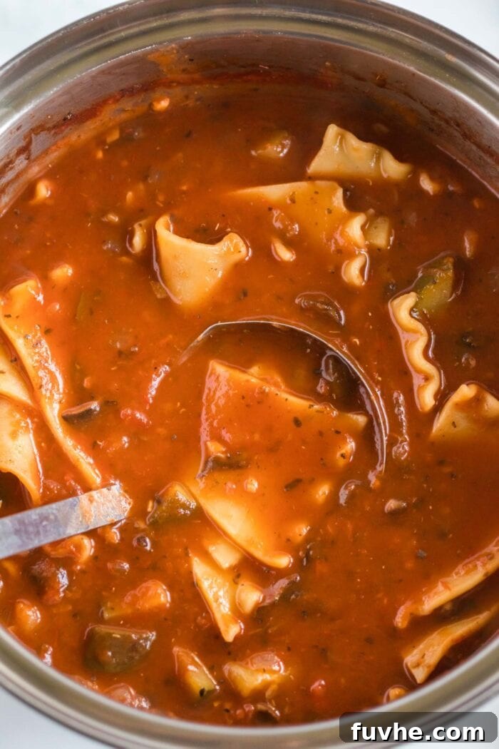 A ladle stirring a large pot of bubbling tomato lasagna soup, with broken lasagna noodles cooking to perfection.