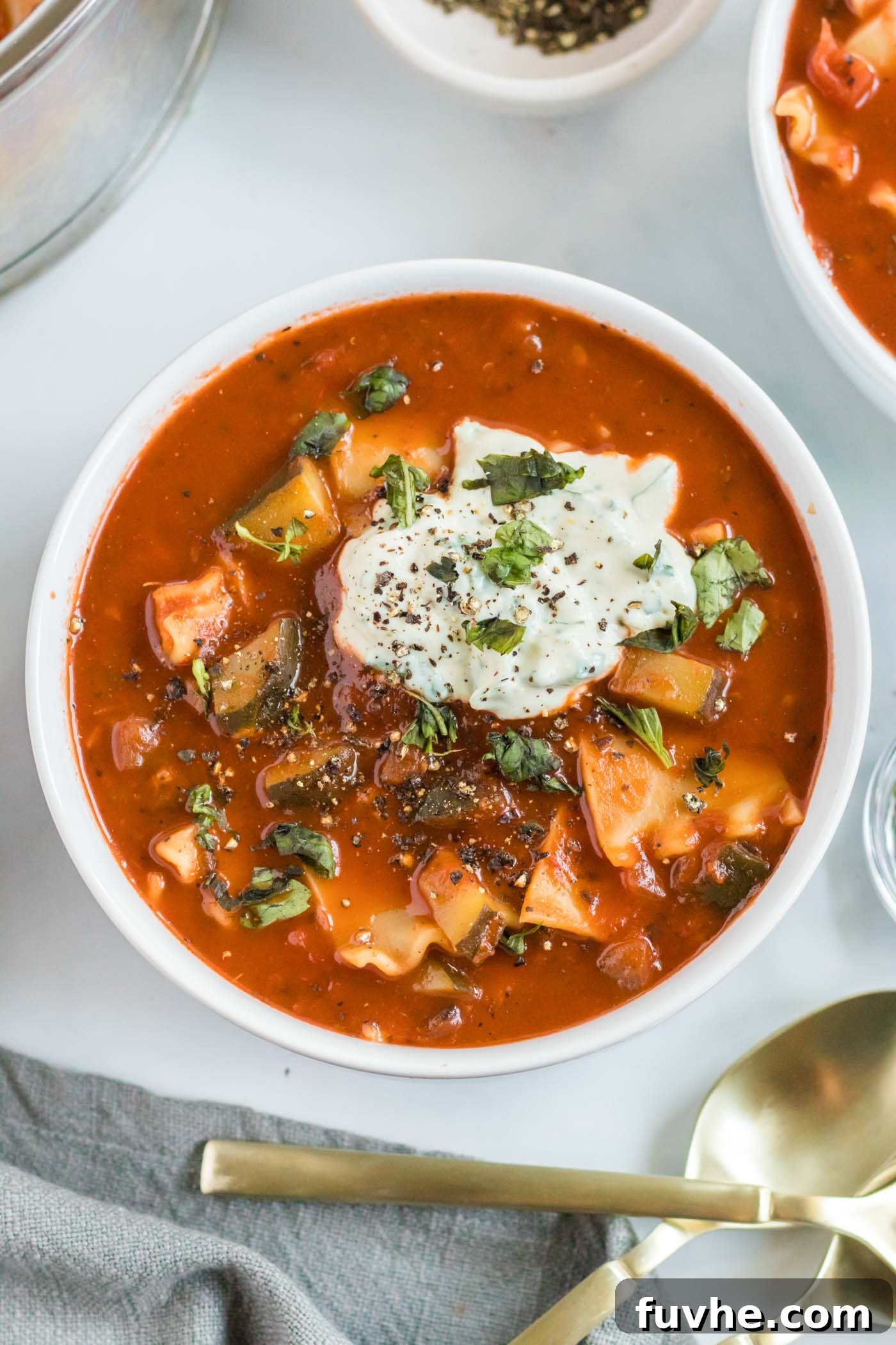 A beautifully presented bowl of vegan lasagna soup, adorned with fresh basil leaves, cracked black pepper, and a generous swirl of creamy tofu ricotta on top.