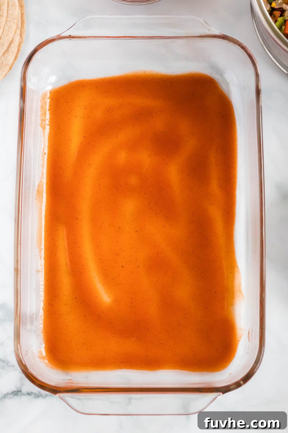 A clear glass baking dish with a thin, even layer of rich red enchilada sauce spread across its bottom, ready for the rolled tortillas.