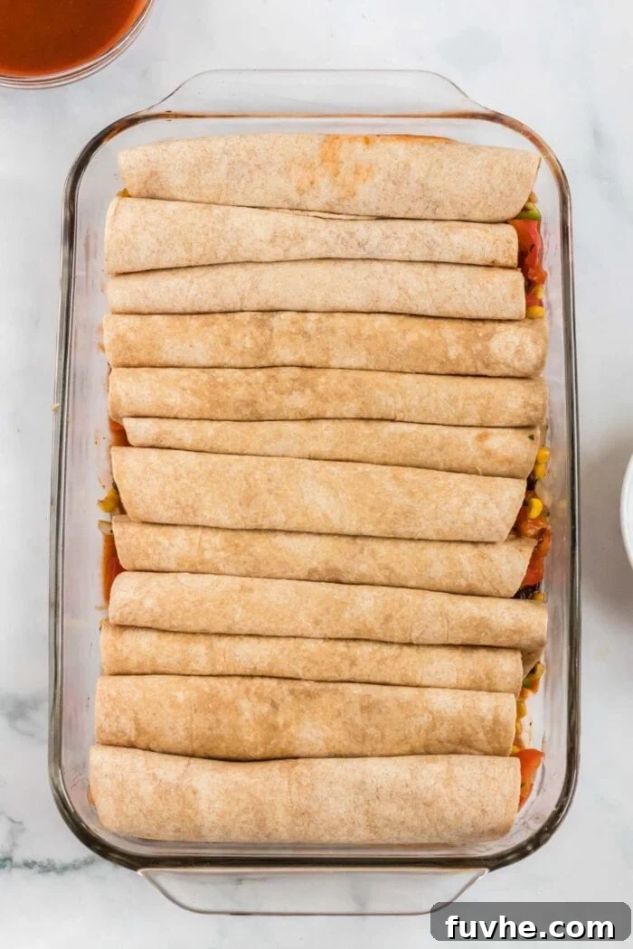 Several neatly rolled enchiladas, filled with black beans and corn, arranged in a single, tight line inside a glass baking dish, ready for sauce and baking.