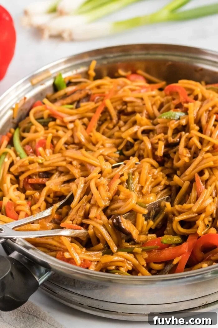 Close up of vegetable lo mein cooking in a large frying pan with a pair of tons in the pan, showing rich sauce and vibrant ingredients.