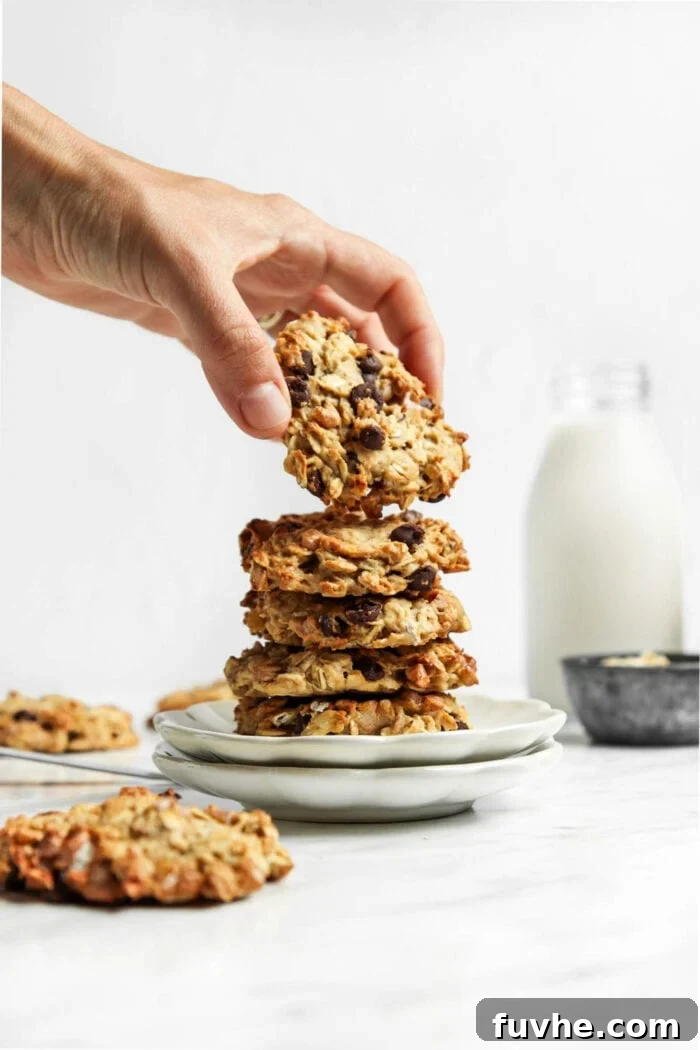 Hand picking a chocolate chip cookie from a stack of freshly baked cookies on a white plate, showcasing their inviting appearance.
