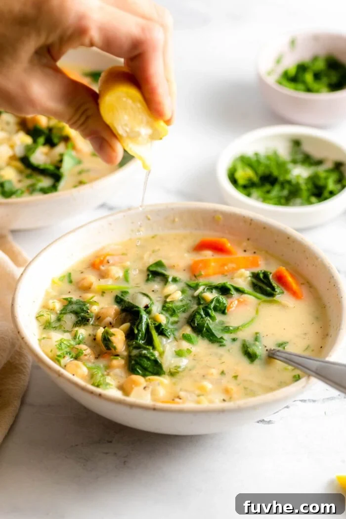 Hand squeezing a lemon wedge over a bowl of lemon chickpea orzo soup with spinach and carrots.