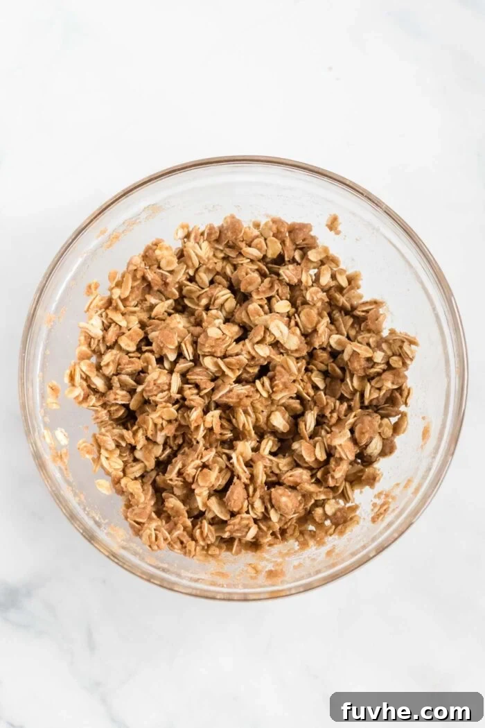 A close-up shot of rolled oats and whole wheat flour being mixed with maple syrup and melted coconut oil in a bowl, forming the rustic crumble topping for the dessert.
