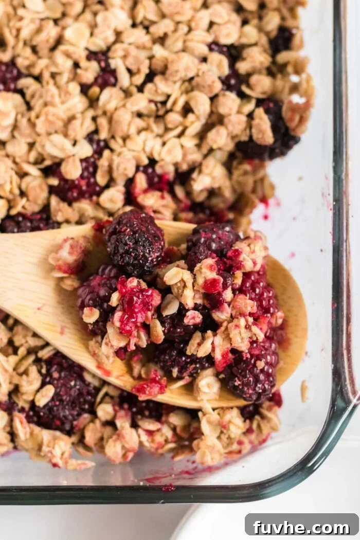 A wooden spoon carefully scooping a portion of warm, baked blackberry crumble from a glass baking dish, highlighting its delicious texture and juicy filling.