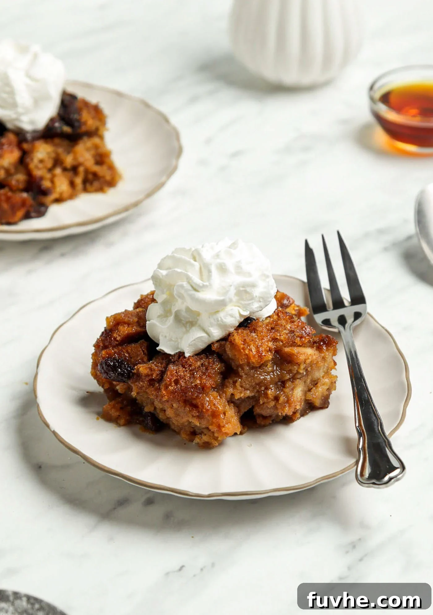 A slice of golden vegan bread pudding topped with fluffy whipped cream and a sprinkle of cinnamon on a plate.