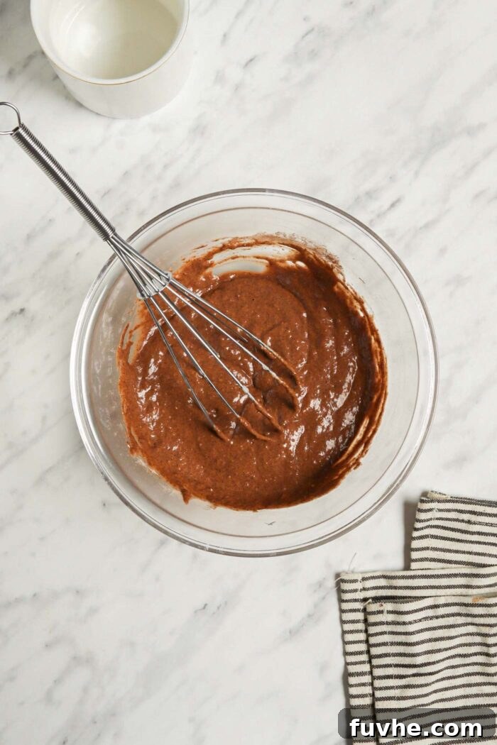 Mugful of Vegan Chocolate Bliss 5 A smooth, rich chocolate cake batter is beautifully mixed together in a clear glass mixing bowl, a whisk resting inside, signaling readiness for baking.