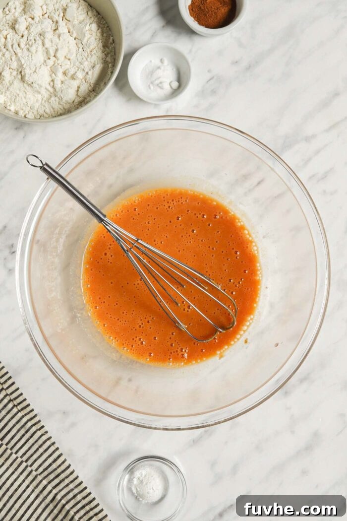 A vibrant orange mixture of pumpkin puree and plant-based milk being smoothly combined in a mixing bowl with a whisk, forming the wet base for the pancake batter.