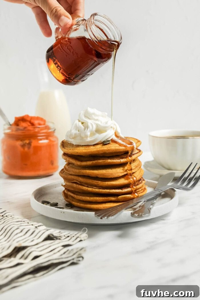 A hand gracefully pouring rich amber maple syrup from a small jar over a towering stack of golden pumpkin pancakes, beautifully adorned with a cloud of vegan whipped cream.
