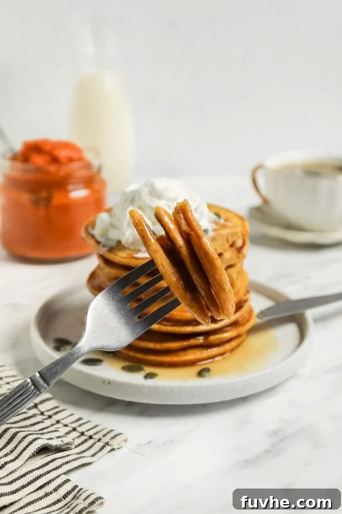 A close-up shot of a fork lifting a piece of perfectly cooked vegan pumpkin pancake, with a stack of more pancakes in the soft focus background, highlighting its fluffy interior.