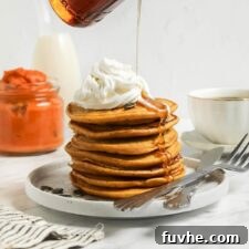 A stack of golden vegan pumpkin pancakes, adorned with vegan whipped cream and a generous pour of maple syrup, captured from an appealing overhead angle.