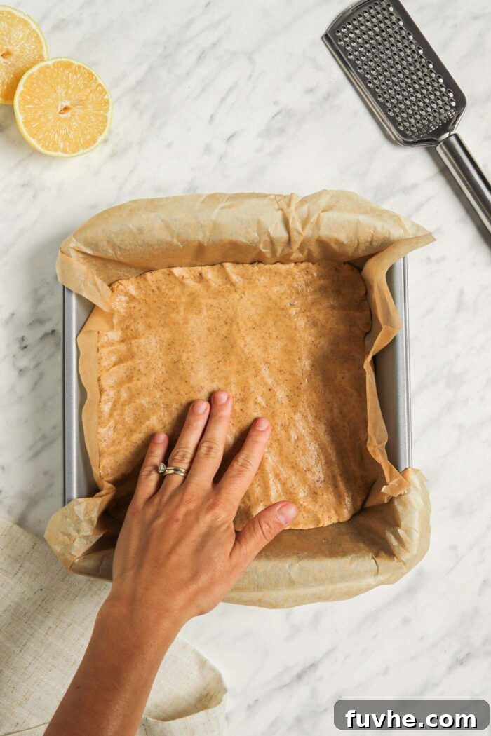A hand pressing raw shortbread dough into a parchment paper-lined square baking dish, meticulously creating an even base.
