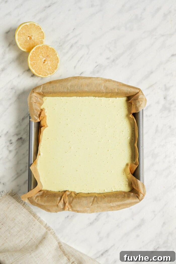 Overhead view of an unbaked vegan lemon bar in a square baking pan lined with parchment paper, with fresh lemon slices placed aesthetically beside the pan.