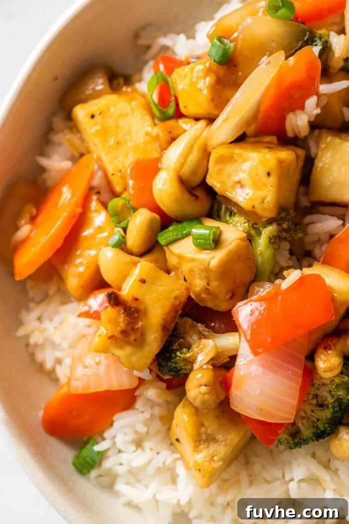 Close up of a tofu stir fry with vegetables and cashews over rice in a bowl.