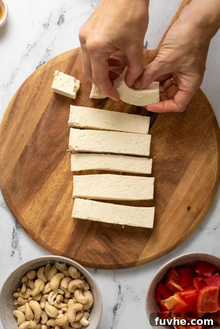 Strips of tofu on a cutting board. Two hands breaking the strips into small cubes.