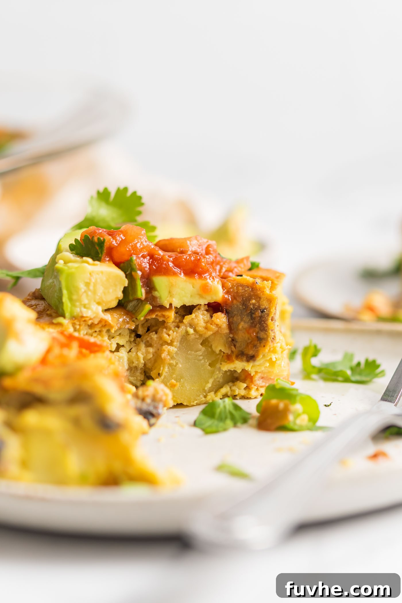 A perfectly baked slice of vegan eggless casserole, showcasing tender chunks of potato and an array of colorful vegetables, beautifully garnished with creamy avocado and fresh salsa.
