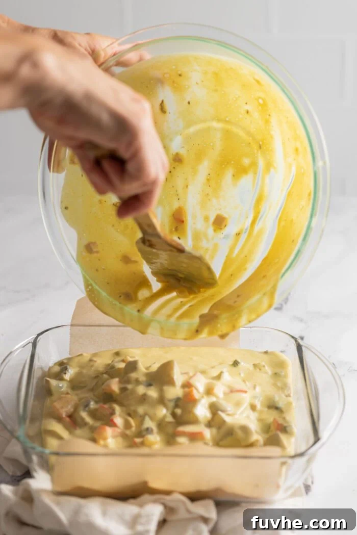 The golden-yellow vegan egg and potato casserole batter being carefully poured from a mixing bowl into a square glass baking pan, ready to be spread evenly and baked.