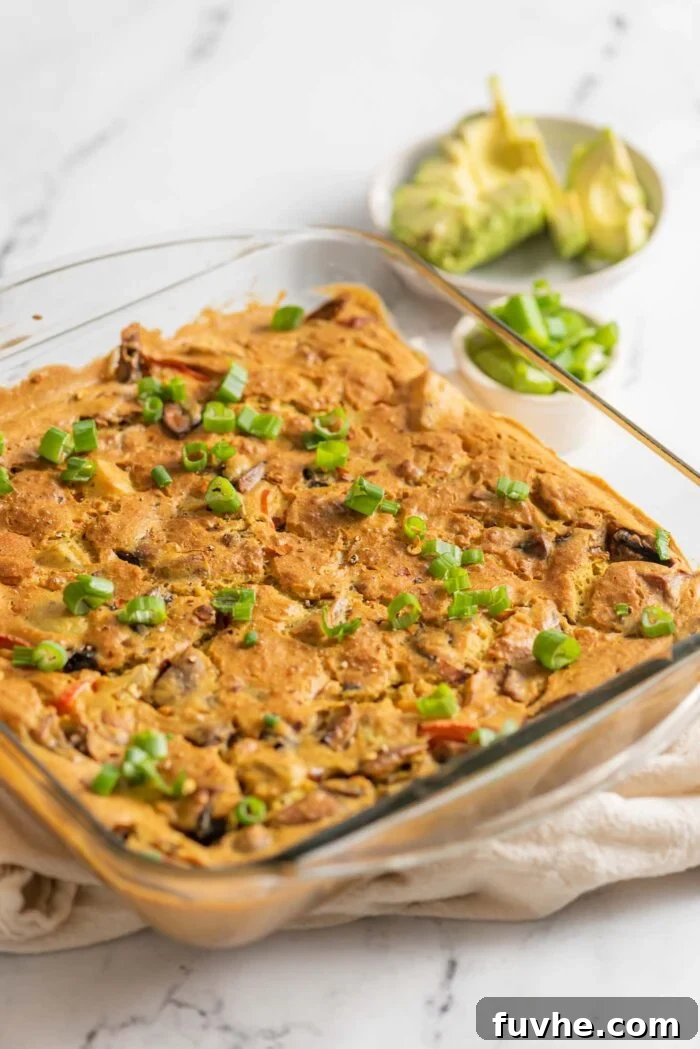 A golden-brown baked vegan potato egg casserole rests perfectly in a glass baking dish, with a small bowl of fresh, sliced avocado visible in the soft background, hinting at delicious toppings.