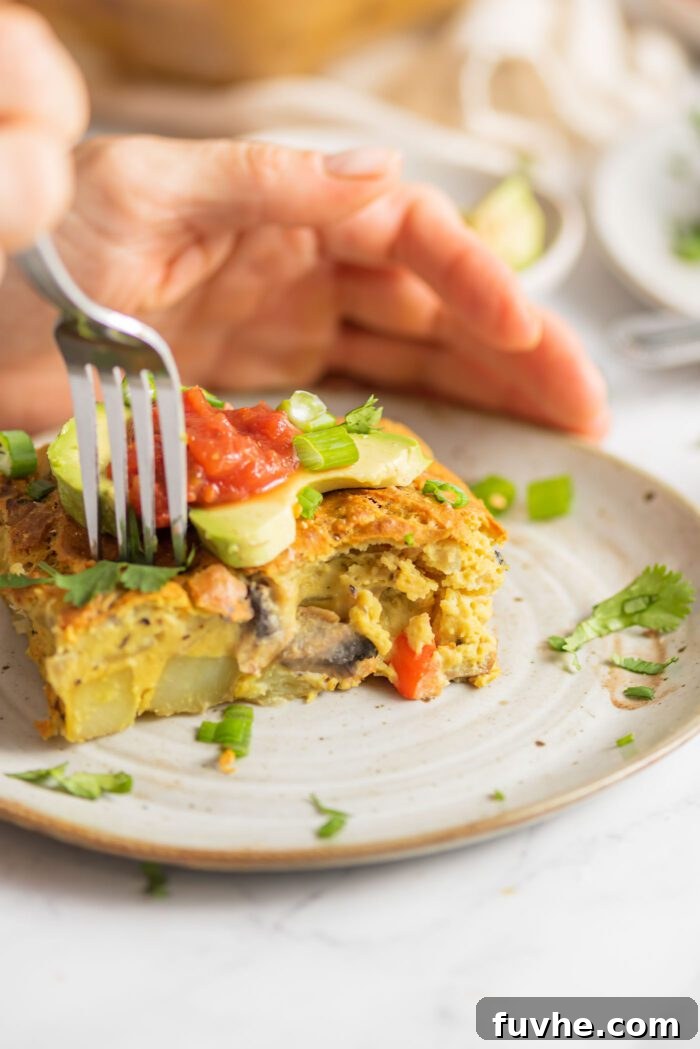 A delectable slice of vegan breakfast casserole, richly filled with potato and vegetables, is topped with creamy avocado and fresh salsa. A hand with a fork reaches in to take a bite, capturing the inviting moment of indulgence.