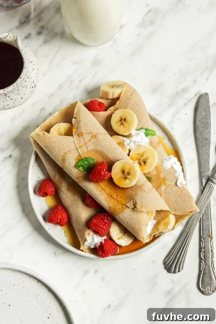 Overhead view of three fluffy vegan buckwheat crepes, generously filled with fresh strawberries, sliced bananas, and a dollop of creamy whipped topping, elegantly arranged on a white plate, ready to be enjoyed as a delightful and healthy meal.